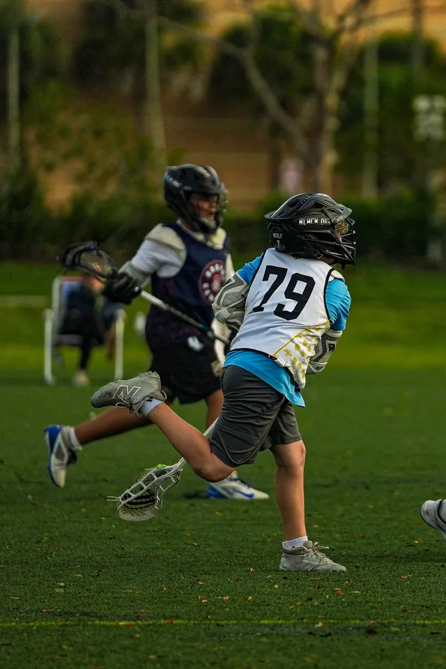 Two helmeted Redhawks boys run with their sticks on the grass in the evening